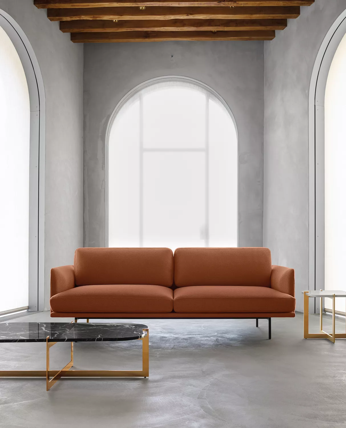 Divano in pelle cognac sotto una finestra ad arco, con tavolini in marmo e ottone, in una hall dai muri in calce