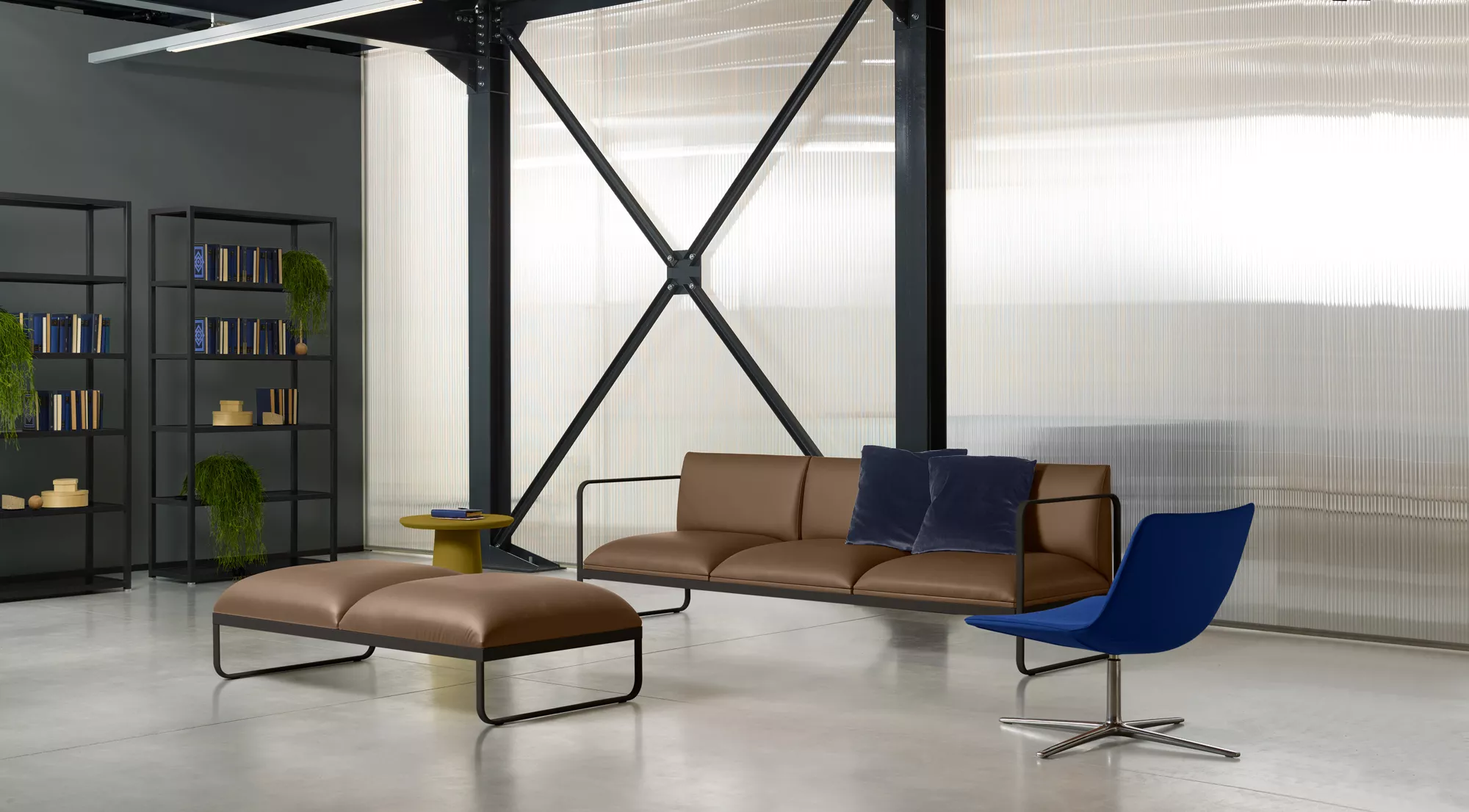 Divano in pelle marrone con telaio metallico a vista, pouf coordinato e poltroncina blu, in un'area comune