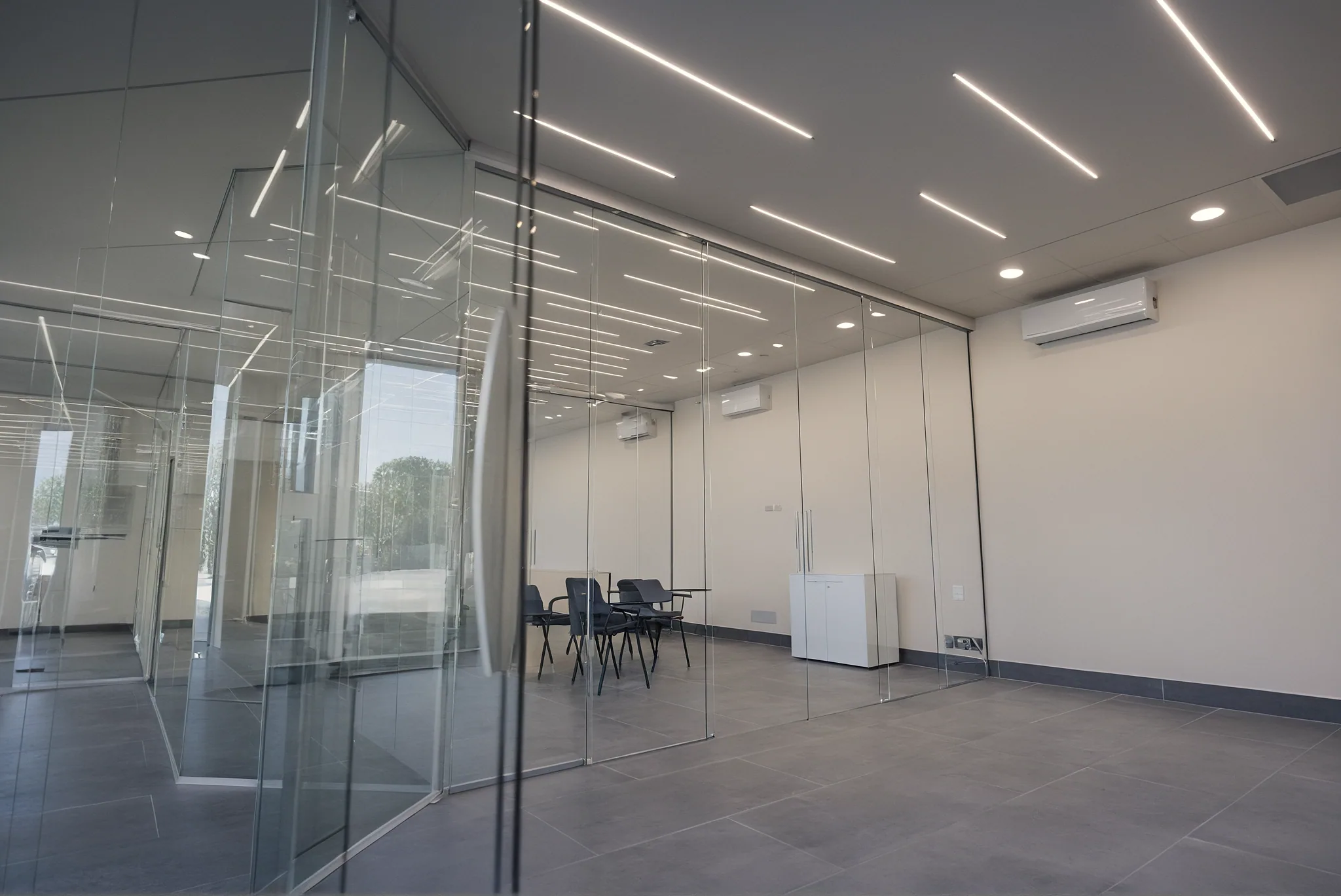 Aula chiusa da pareti in vetro con porte scorrevoli appese a un binario continuo, vista dal corridoio; dentro, sedute nere con tavoletta e un mobile basso