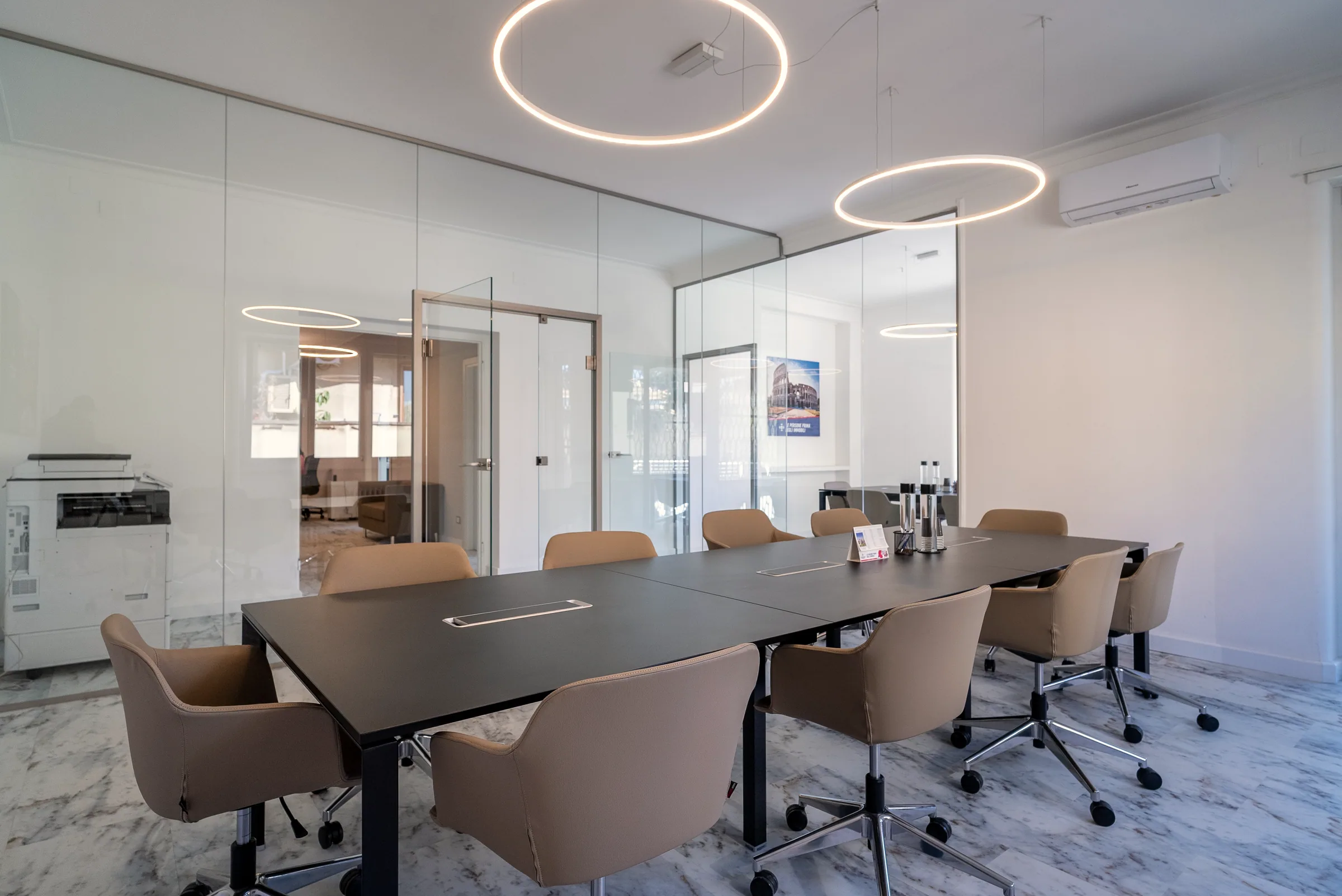 Sala riunioni luminosa con grande tavolo scuro, sedute beige, pareti vetrate e lampade circolari sospese.