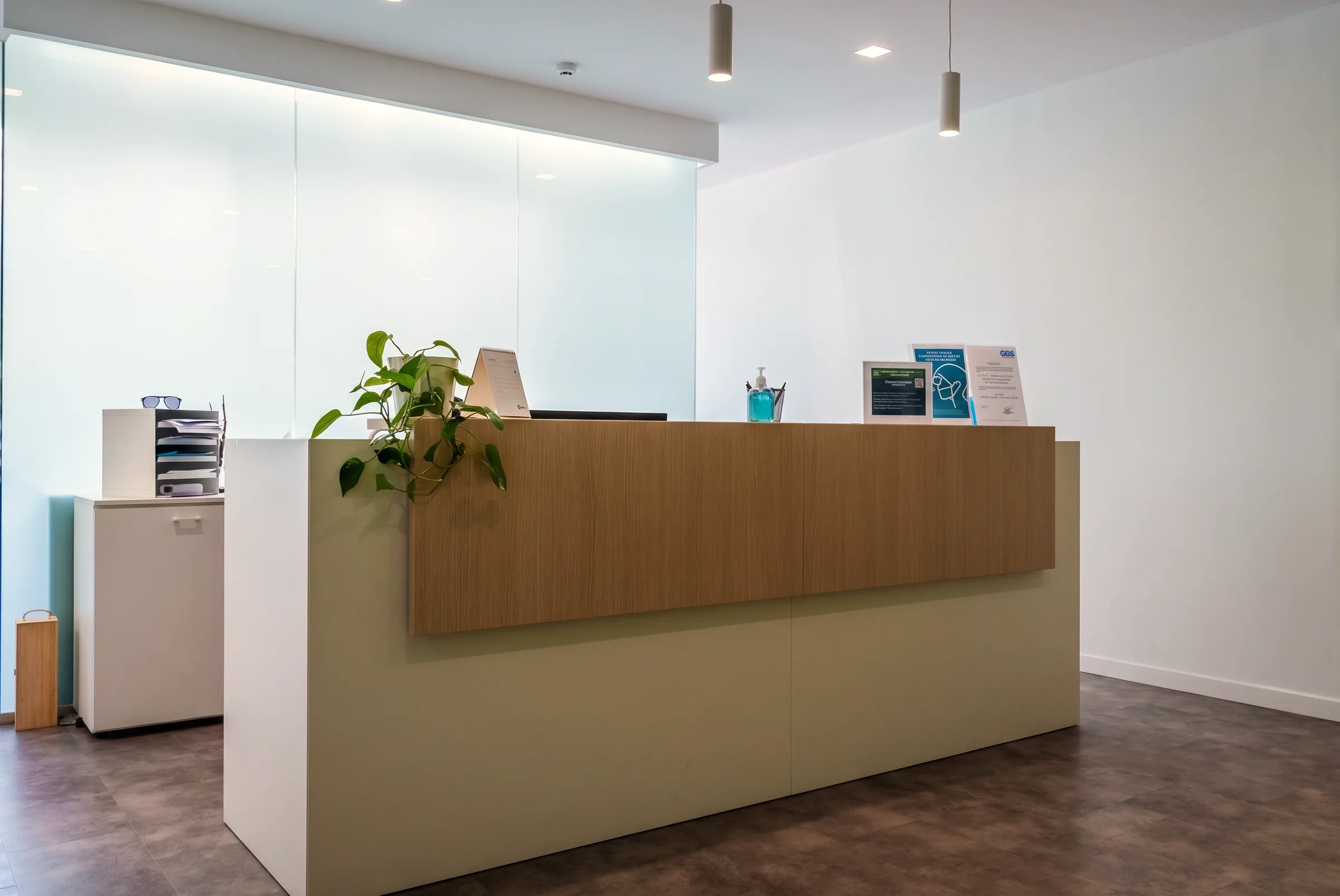 Reception installata in ufficio: bancone verde salvia con fascia in rovere e fianco bianco, parete vetrata retroilluminata, pianta e cartelli informativi.
