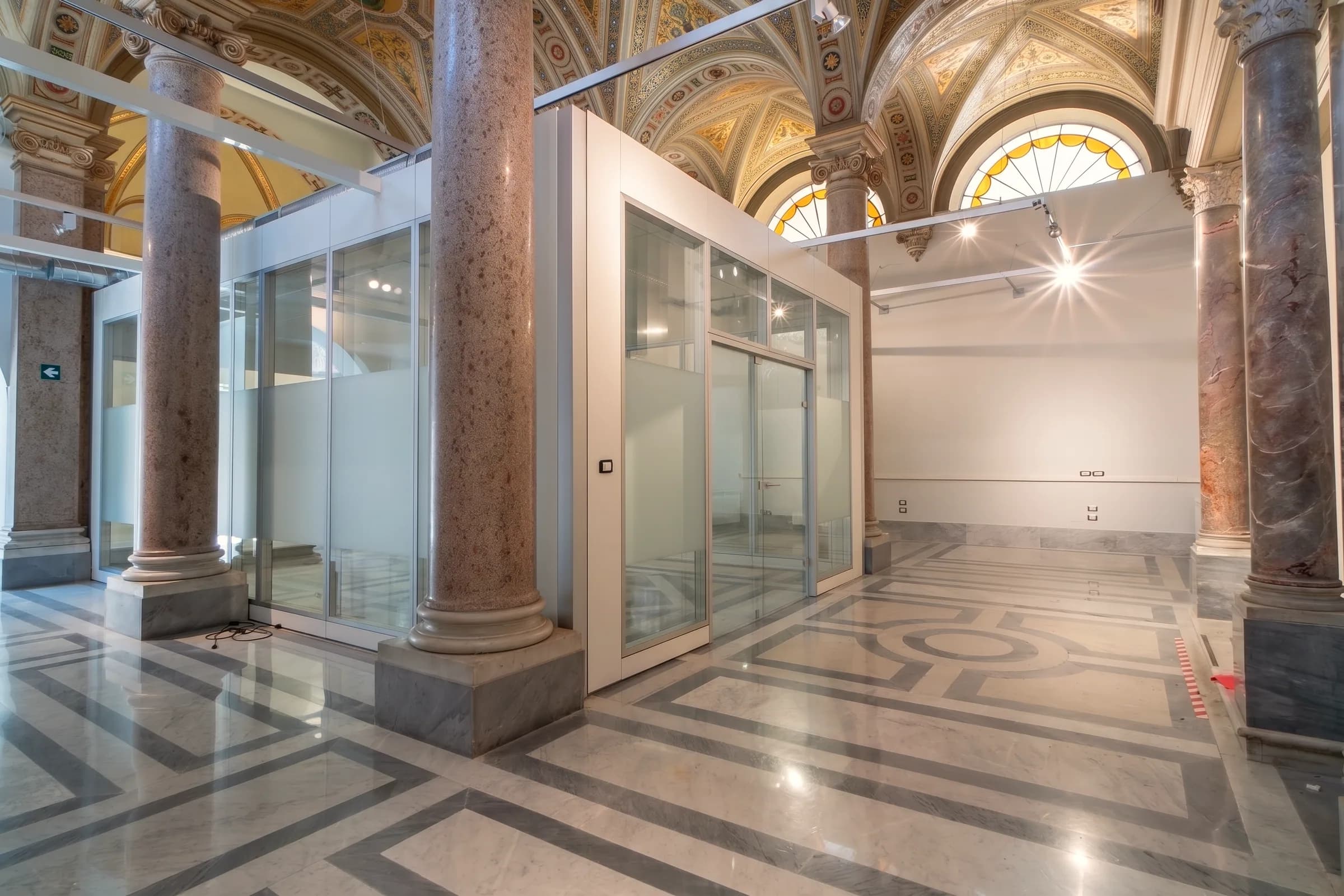 Sala vuota delimitata da pareti in vetro, con alte finestre ad arco e pavimento in marmo con inserto rosso