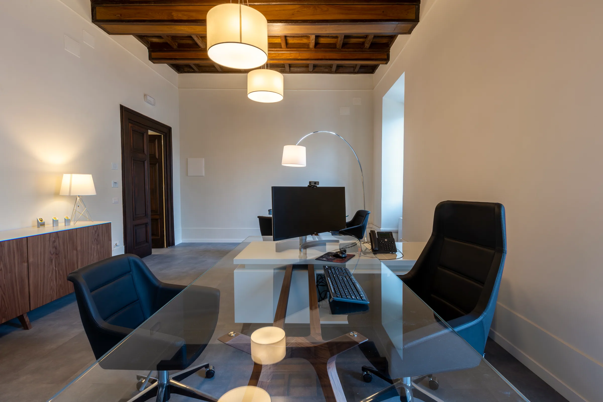 Ufficio direzionale consegnato sotto un soffitto a travi in legno: scrivania bianca con piano in vetro, poltrone in pelle nera e credenza in legno