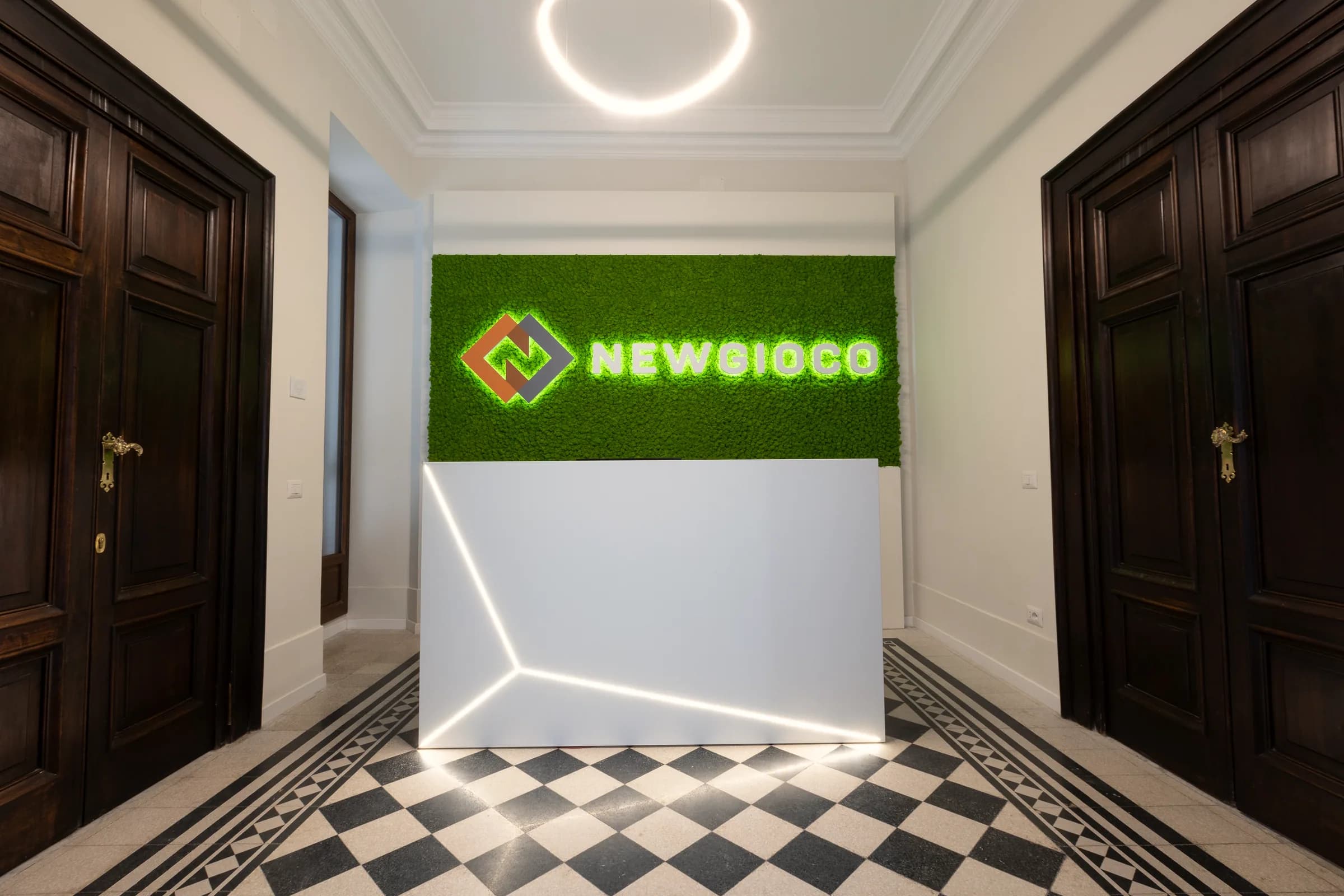 Reception con parete vegetale verde, logo Newgioco retroilluminato, bancone bianco percorso da tagli luminosi e pavimento in cementine bianche e nere a scacchi