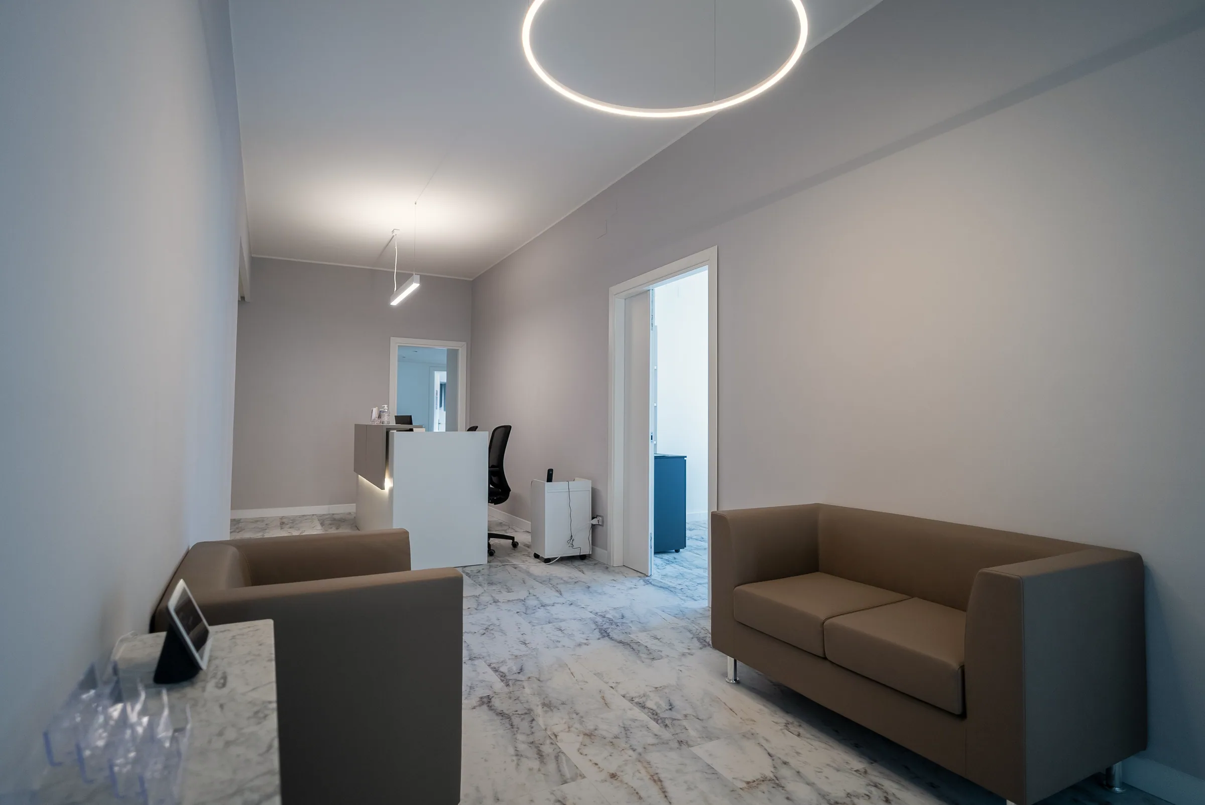 Area di attesa con due divanetti beige, lampada ad anello a soffitto e, in fondo, il fianco del bancone della reception